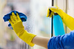 Two hands wearing yellow latex gloves spraying cleaning solution onto a window.