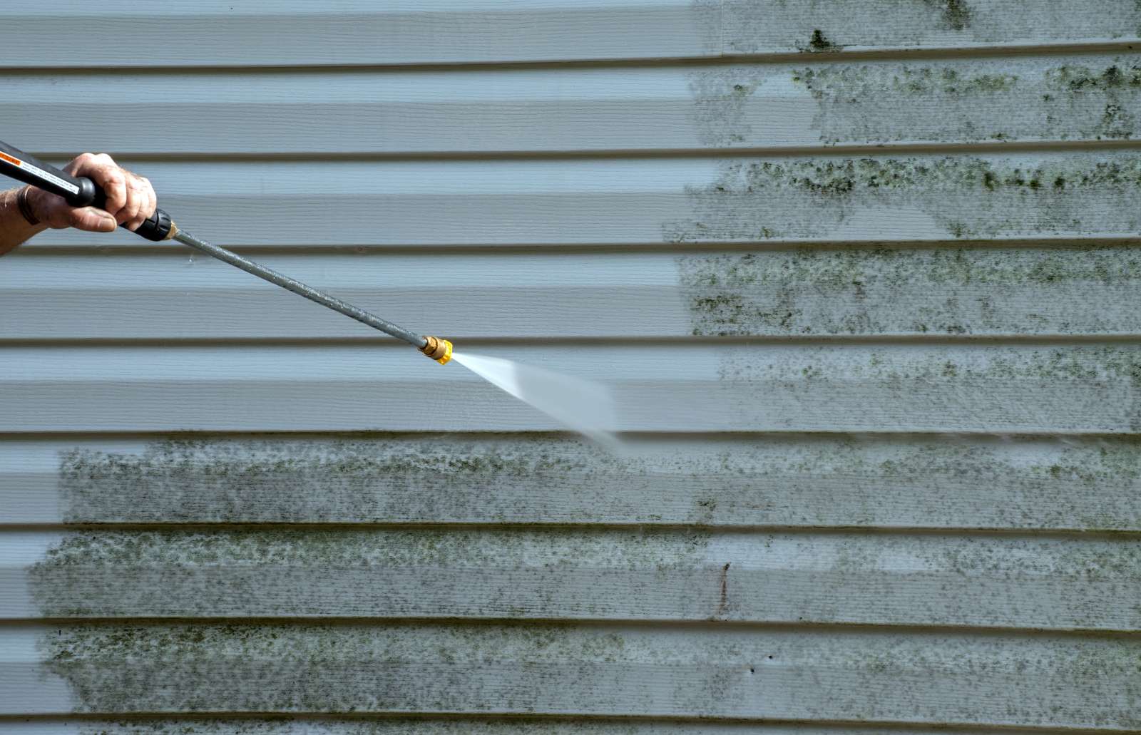 A pressure washing nozzle spraying water at a home's siding, half of which is clean while the other half is dirty.
