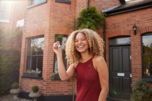 A smiling woman holding up a house key in front of a large red brick home.